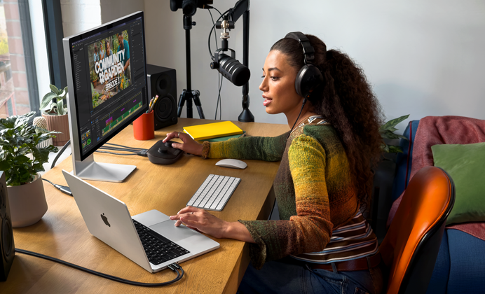 Una mujer en su casa trabaja en su mesa con un MacBook Pro conectado a un monitor externo, lleva auriculares y habla por un micrófono
