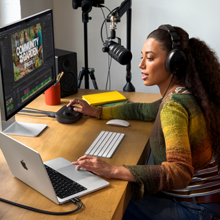 Una mujer en su casa trabaja en su mesa con un MacBook Pro conectado a un monitor externo, lleva auriculares y habla por un micrófono