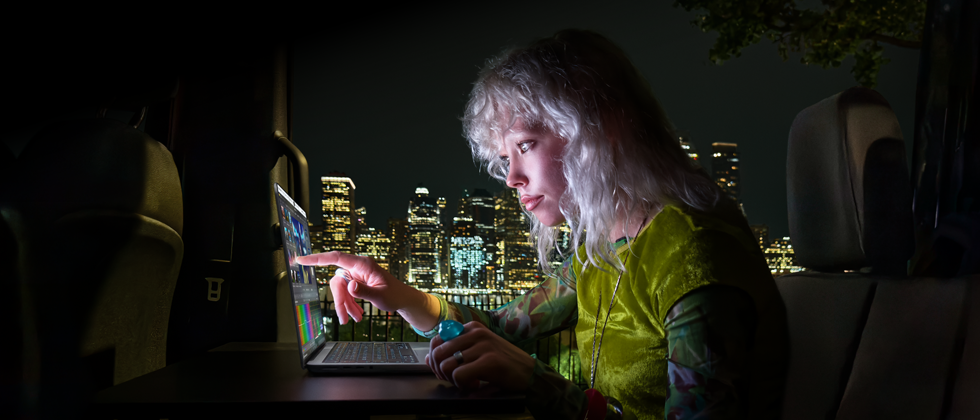 Una persona sentada en el asiento trasero de un furgón, la puerta del pasajero abierta muestra el horizonte de una ciudad por la noche, MacBook Pro con una pantalla PROLED XDR sobre una mesa de trabajo, la persona está iluminada por la pantalla mientras utiliza el control táctil para editar un vídeo en DaVinci Resolve Studio
